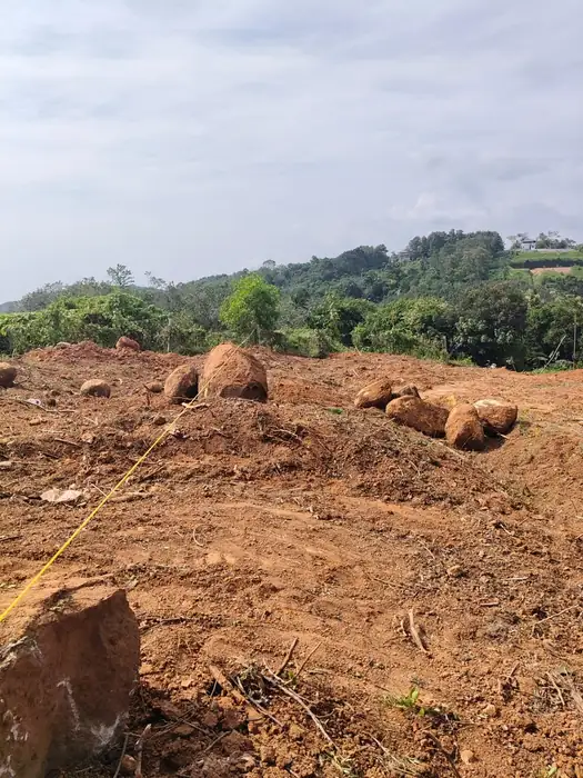 Cleared plot on sloping ground with boulders and a setting-out line, wooded hills beyond